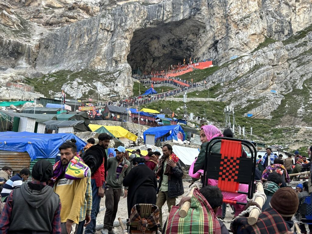 अमरनाथ यात्रा के दौरान खच्चर और पालकी से पहाड़ी रास्तों पर चढ़ते श्रद्धालु, सामने बर्फ से ढकी चोटी, एक तरफ़ टूटे रास्ते पर गिरती युवती, दूसरी ओर बाबा बर्फानी की गुफा के दर्शन करते श्रद्धालु — थके, डरे, लेकिन संतुष्ट। वातावरण में बर्फ, कीचड़ और आस्था का समावेश।